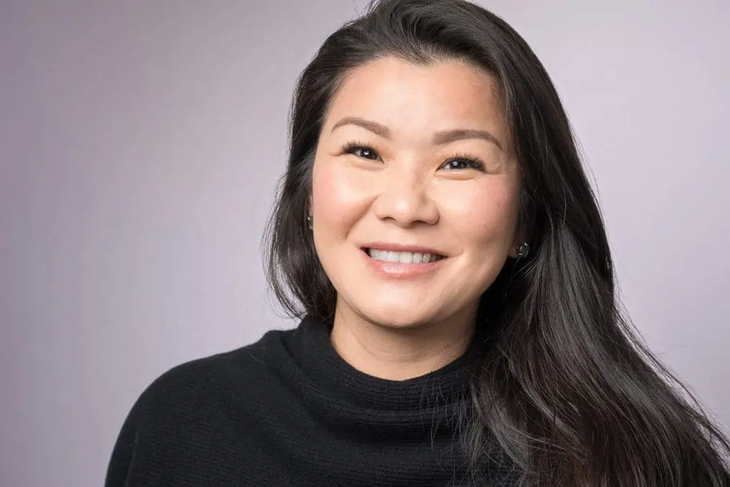 A woman with long dark hair and a black sweater smiles at the camera against a light gray background. Photography by Jack Simpson