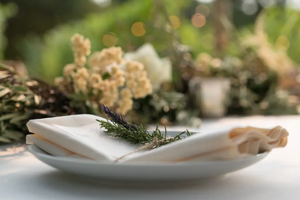 A white plate holds a neatly folded napkin tied with twine and topped with a sprig of lavender and rosemary. Shot by Napa event photographer Jack Simpson, soft-focus flowers and greenery are visible in the background.
