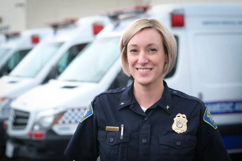 A person in a police uniform smiles at the camera. They stand in front of a row of parked ambulances. The uniform includes a badge and shoulder insignia. The background is slightly out of focus.