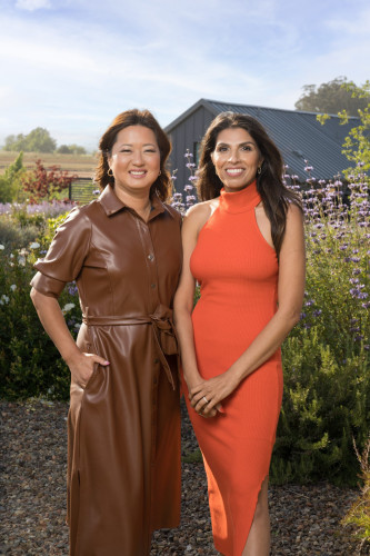 Two women stand outdoors in a garden. One wears a brown leather dress, and the other wears an orange sleeveless dress. Behind them is a dark building and colorful flowers. The sky is clear and sunny.