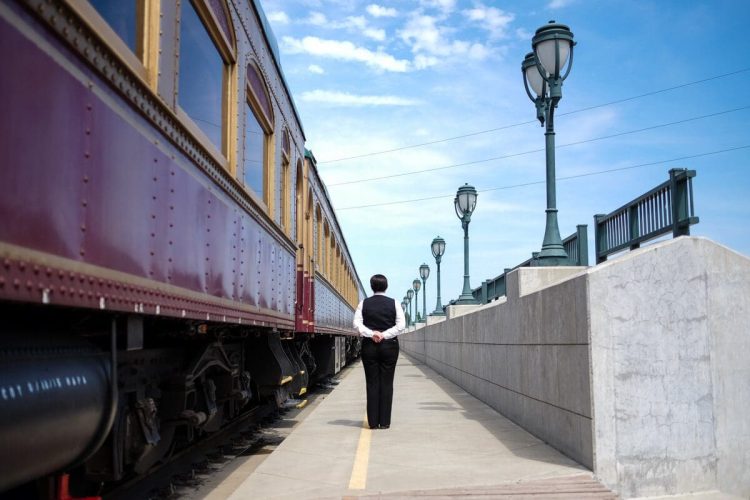 A person in uniform stands on a platform next to a stationary train, with burgundy-colored carriages. They have their hands clasped behind their back. Streetlights and a clear blue sky are visible in the background.