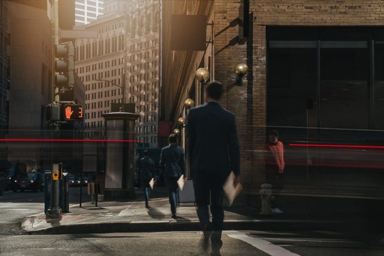 Business people walking across a city street under tall buildings. A crosswalk sign shows a red hand, and a moving light trail blurs past. Its daytime with a mix of natural and artificial light.
