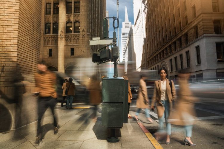 A city street scene with blurred pedestrians walking at a crosswalk on a sunny day, surrounded by tall buildings and a traffic signal in the center foreground. Photography by Jack Simpson
