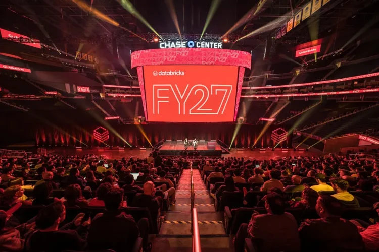 An elevated, wide view of the Chase Center stadium interior during a large corporate event, showing hundreds of seated attendees facing a stage and the massive center-hung screen displaying "Databricks FY27."